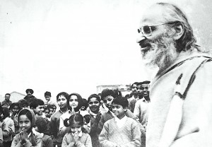 Black and white photo of smiling Indian man with a beard in profile. He is surrounded by young Indian children and a few adults