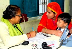 Three Indian people - a woman doctor, a mother wearing a red head covering, and a small boy - discussing health concerns