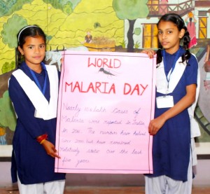 Two Indian school girls in blue and white salwar kameez uniforms hold up a sign saying 'World Malaria Day'