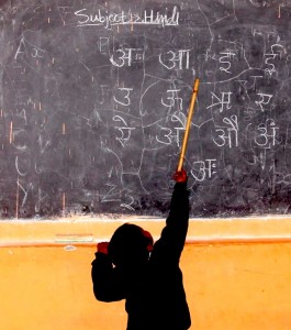 A small Indian boy uses a stick to reach up to point at Hindi letters written in white on a black chalkboard